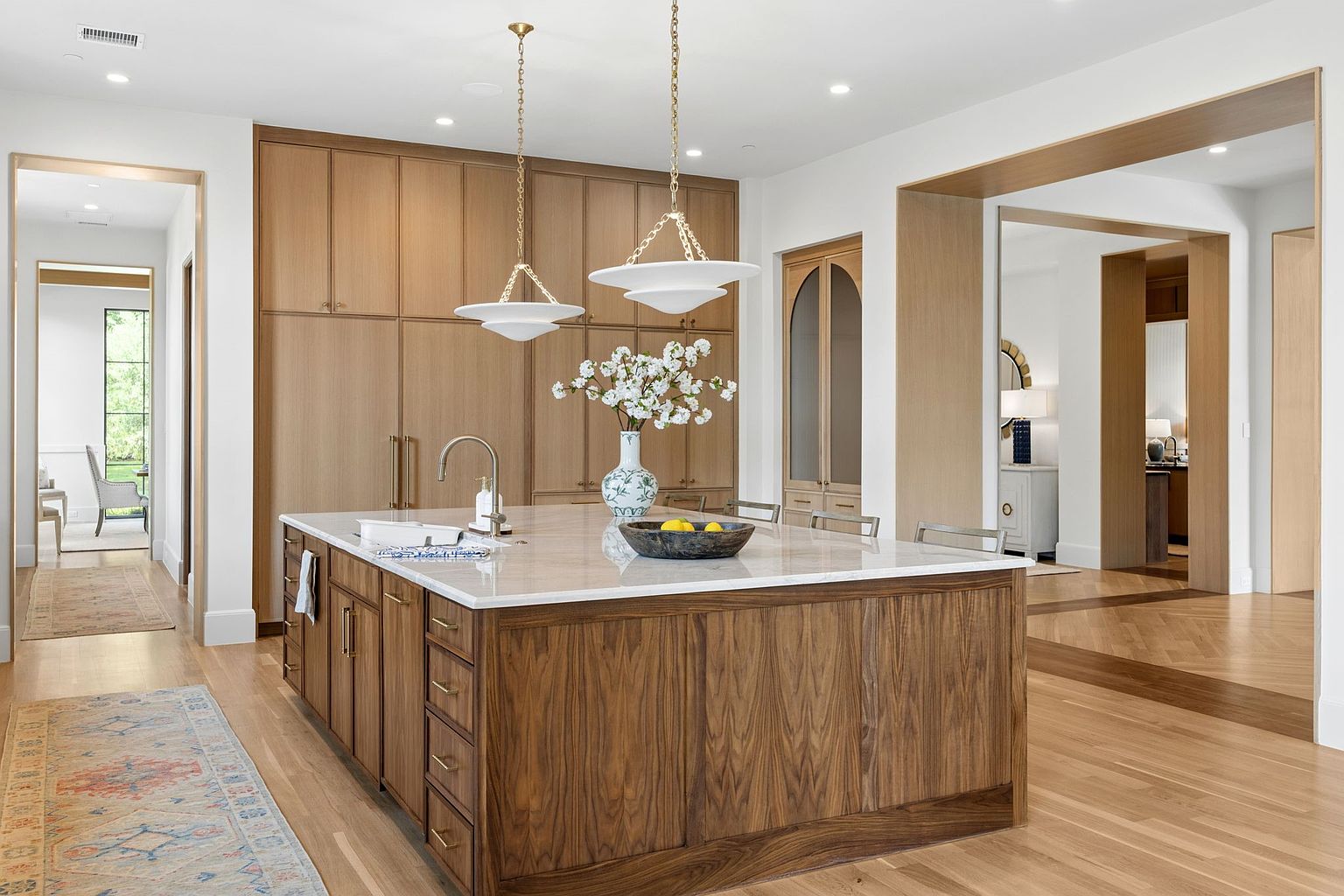 This sophisticated kitchen features a large central island with a stone countertop and dark wood cabinetry, complemented by light wood cabinetry along the back wall. Two elegant pendant lights hang above the island, which is accented by a decorative vase and a bowl of fruit. The space is framed by wide doorways and light wood flooring, creating an open and airy atmosphere with a seamless transition between rooms.