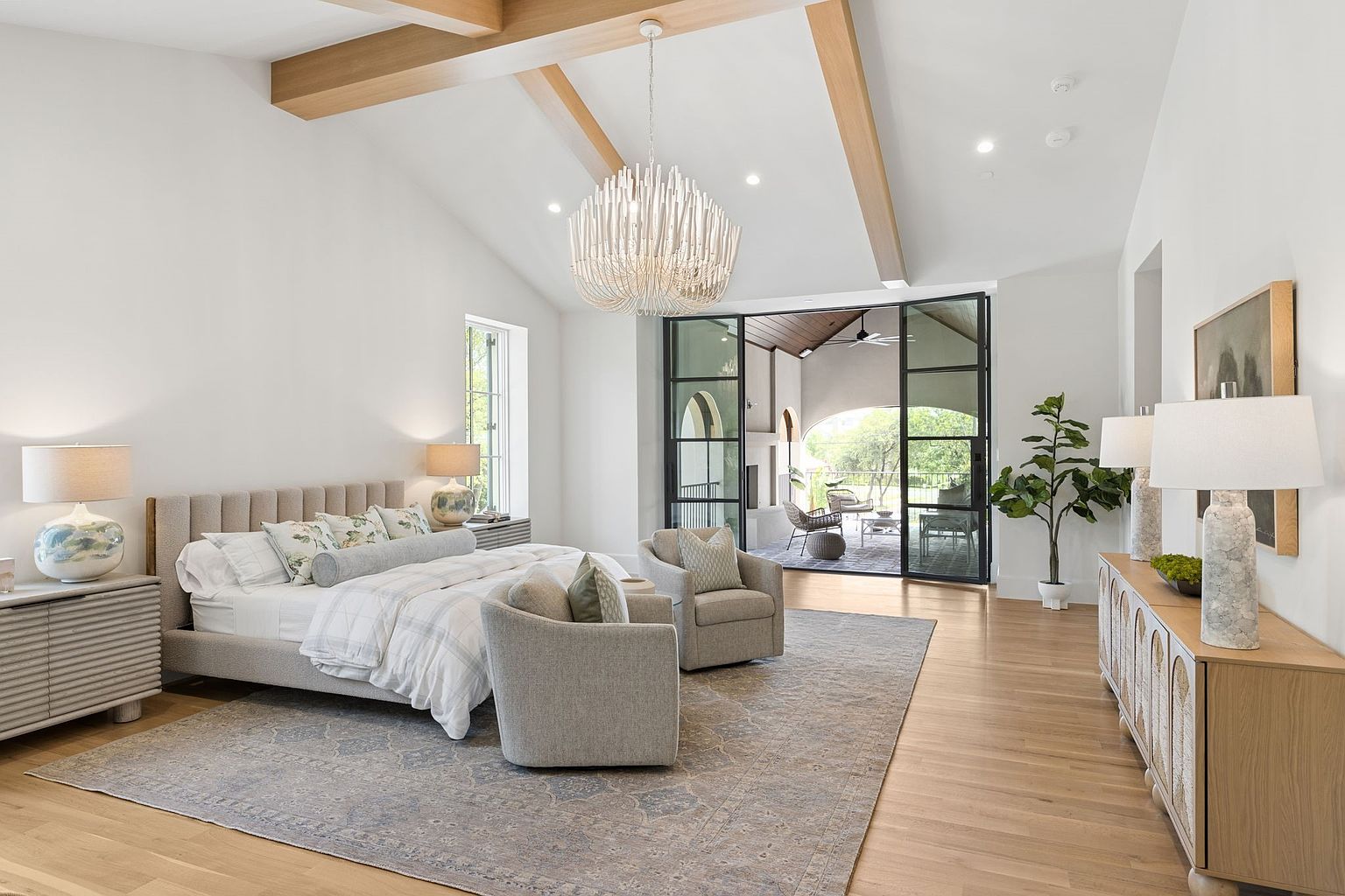 This spacious primary bedroom features a vaulted ceiling with exposed light wood beams and a striking modern chandelier. A large upholstered bed sits centered on a patterned area rug, flanked by two armchairs that create a cozy seating area. Large glass doors open to a covered patio, allowing natural light to flood the room and providing a seamless transition to the outdoor space.