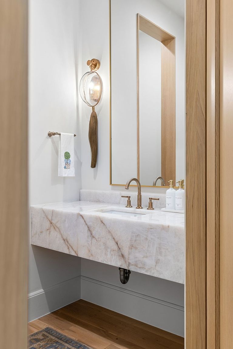 This elegant powder room features a floating vanity topped with a striking, heavily veined stone slab. A tall, thin-framed mirror sits above the vanity, complemented by a unique, sculptural wall sconce with a hanging tassel detail. The space is finished with light wood flooring and a minimalist aesthetic, creating a sophisticated and airy atmosphere.