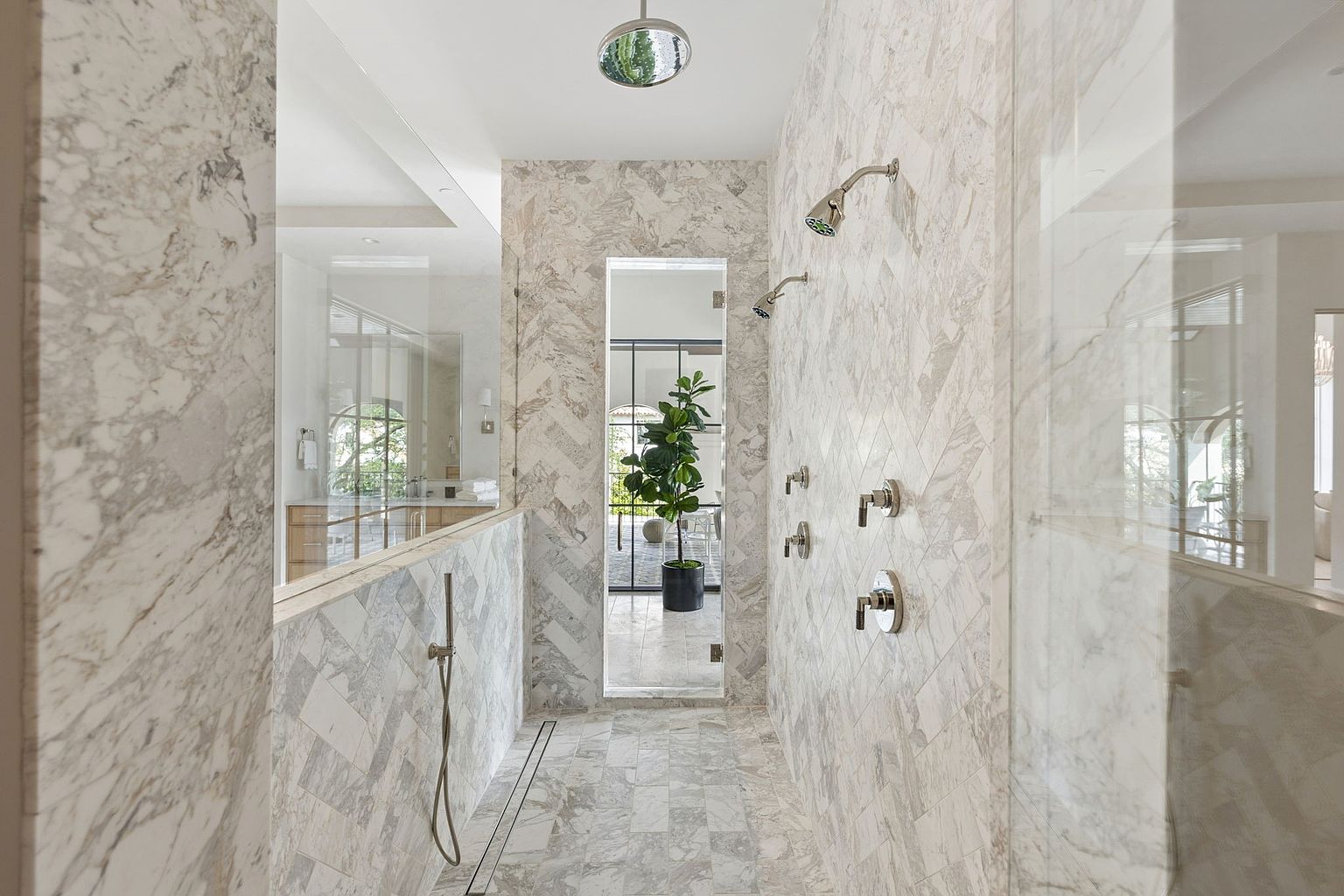 This luxurious walk-in shower features intricate herringbone-patterned stone tile work on the walls and floor, creating a sophisticated and cohesive aesthetic. The space is equipped with dual shower heads, multiple wall-mounted controls, and a handheld sprayer, all finished in a polished metallic tone. A glass partition separates the shower from the adjacent room, while a narrow doorway at the back leads to a bright, open area featuring a potted plant, enhancing the airy and high-end feel of the bathroom.