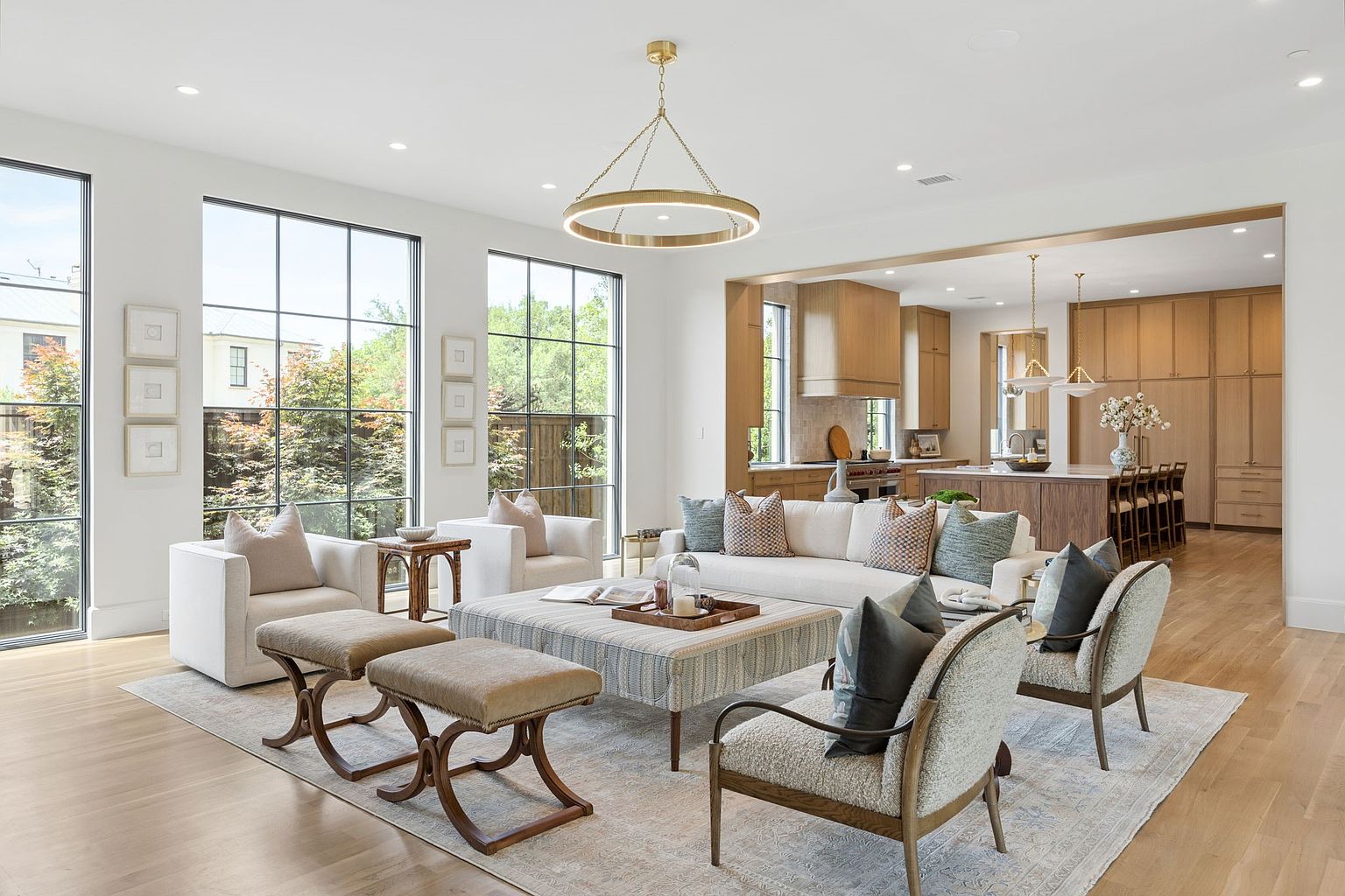 This bright and airy living room features an open-concept layout that flows seamlessly into a spacious kitchen with light wood cabinetry. The space is anchored by a large, patterned ottoman and a neutral sofa, complemented by two armchairs and a pair of upholstered benches. Tall, grid-paneled windows allow for abundant natural light, creating an inviting and sophisticated atmosphere with a modern aesthetic.