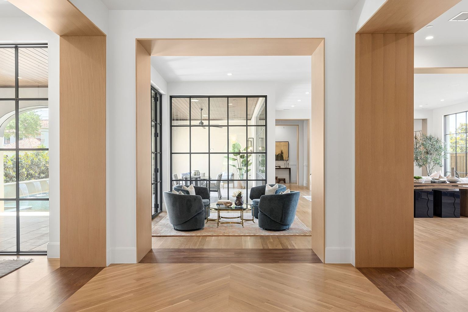 This perspective captures a sophisticated transition space featuring light wood flooring arranged in a chevron pattern and framed by prominent light wood archways. Through a large, black-framed glass partition, a cozy seating area with two blue armchairs and a small coffee table is visible, creating a sense of depth and layered living. The clean, white walls and minimalist design aesthetic emphasize an open, airy, and high-end architectural style.