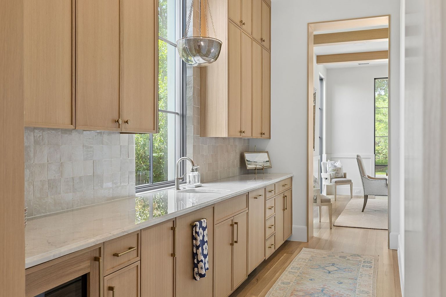This bright and airy butler's pantry or secondary kitchen area features light wood cabinetry with modern brass hardware and a polished stone countertop. A tall window provides natural light, complemented by a decorative pendant light, while a patterned tile backsplash adds texture to the space. The view extends through a doorway into a dining area, showcasing a cohesive, high-end design with light wood flooring throughout.