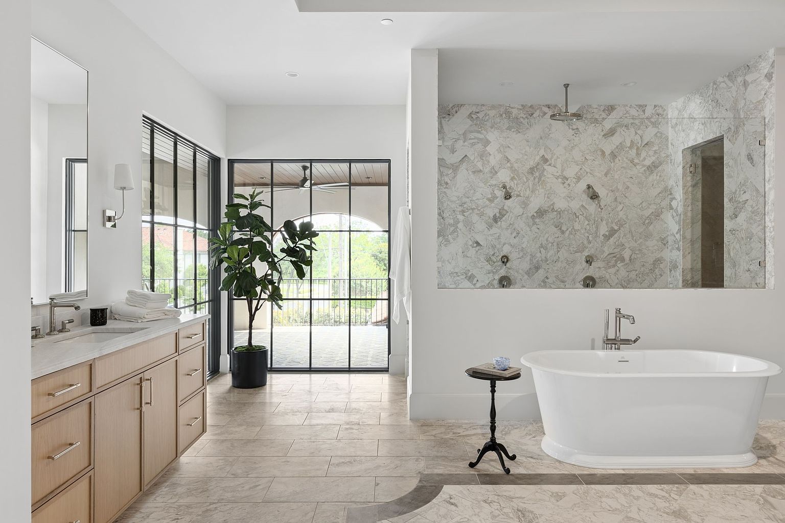 This spacious primary bathroom features a light wood vanity with a stone countertop and a freestanding soaking tub positioned near a large walk-in shower with patterned tile walls. Large, black-framed windows and a matching glass door provide ample natural light and a view of the outdoor patio area. The room is finished with neutral stone flooring, creating a bright, clean, and luxurious atmosphere.