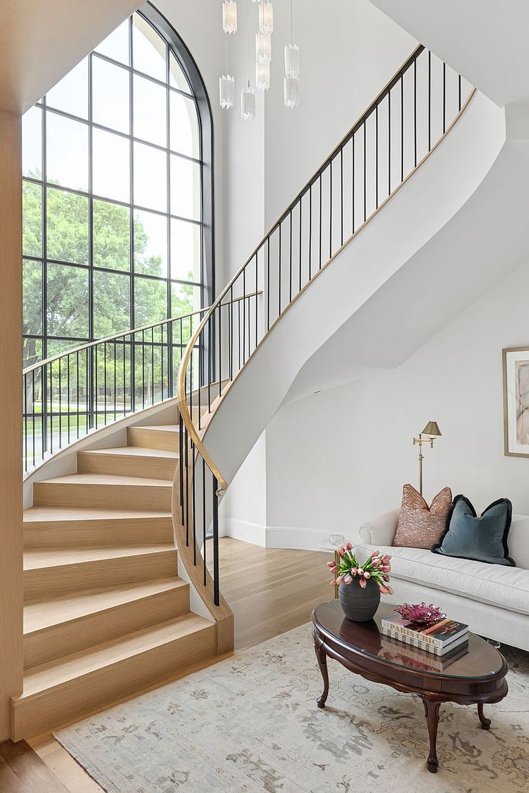 This elegant entryway features a sweeping curved staircase with light wood treads and a sleek black metal railing topped with a warm-toned handrail. A massive arched window floods the space with natural light, highlighting the airy, neutral-toned walls and the sophisticated chandelier hanging above. To the side, a cozy seating area is furnished with a light-colored sofa, plush pillows, and a dark wood coffee table, creating a welcoming and refined atmosphere.