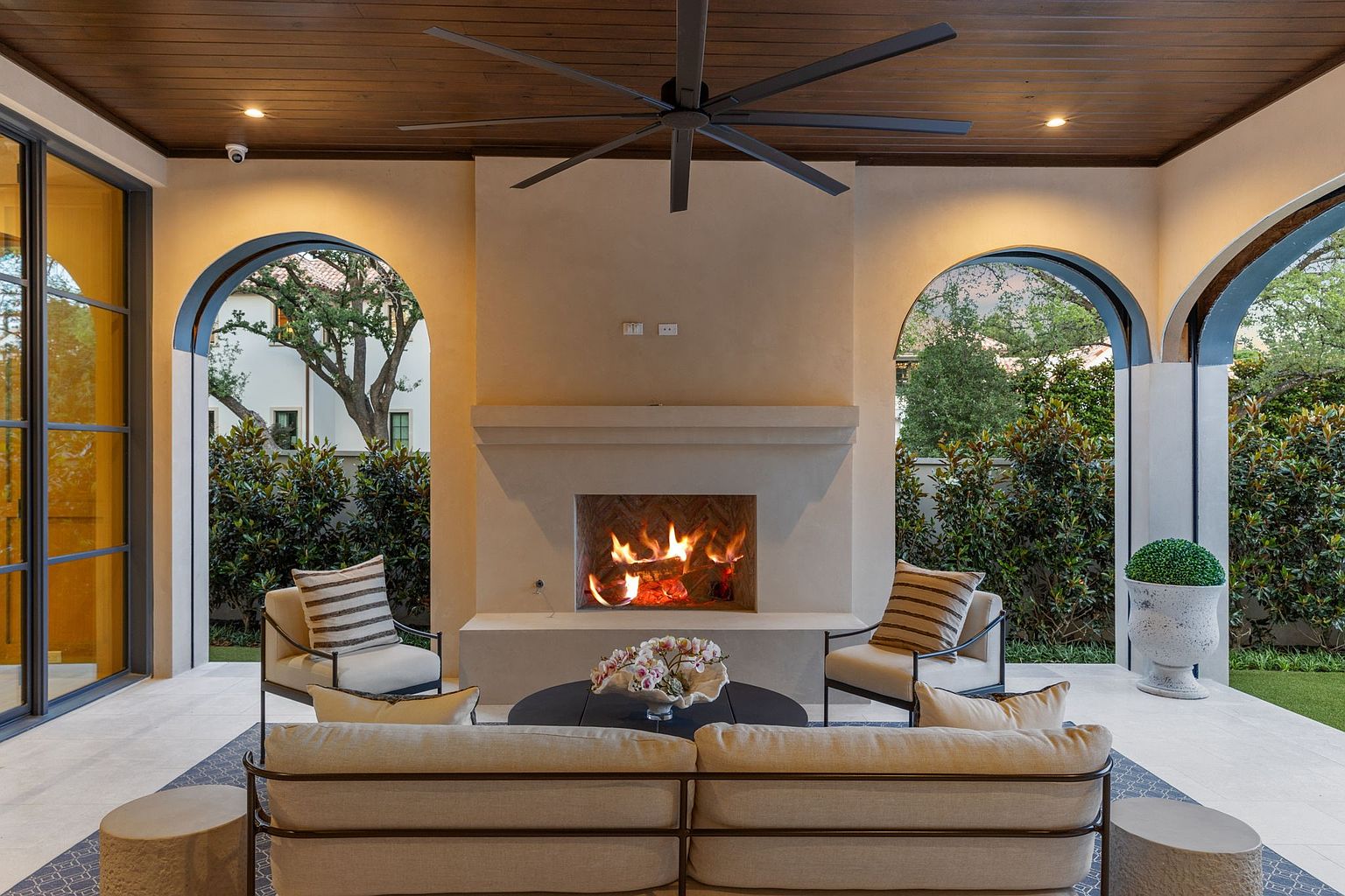 This elegant covered patio features a central fireplace with a light-toned surround and a herringbone-patterned firebox, creating a cozy focal point. The space is furnished with a comfortable sofa and two armchairs arranged on a patterned rug, all set beneath a dark wood-paneled ceiling with a large overhead fan. Arched openings frame views of the lush greenery outside, offering a seamless transition between indoor comfort and the outdoor landscape.
