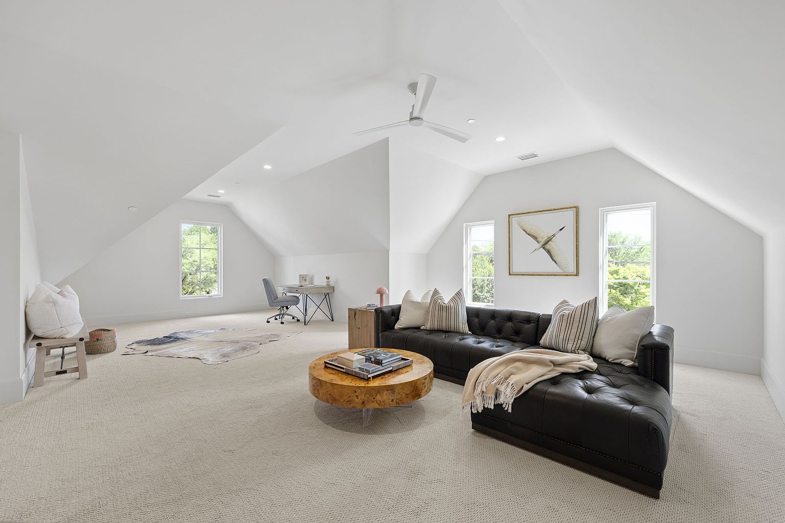 This spacious, light-filled attic room features a vaulted ceiling and a neutral color palette, creating an airy and versatile living space. The area is furnished with a dark leather tufted sectional sofa, a round wooden coffee table, and a dedicated office nook with a desk and chair in the background. Large windows on multiple walls allow for ample natural light, while a patterned area rug and decorative pillows add texture and warmth to the room.