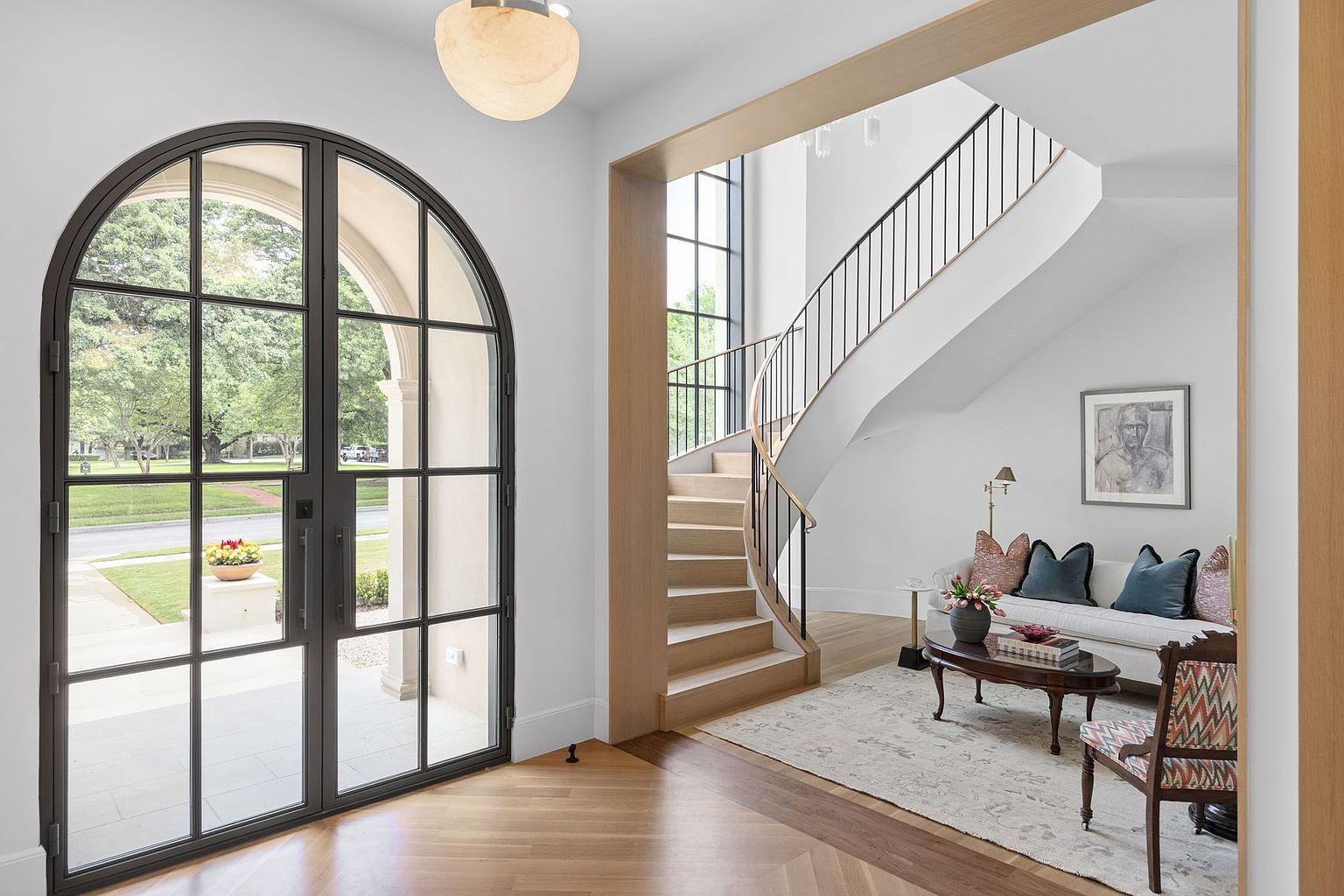 This elegant entryway features a grand, curved staircase with light wood treads and a sleek black metal railing. A large, arched metal-framed glass door provides a bright, welcoming view of the front yard, while a cozy seating area with a sofa and patterned chair sits tucked under the staircase. The space is finished with light wood flooring arranged in a herringbone pattern and a large, spherical pendant light, creating a sophisticated and airy atmosphere.