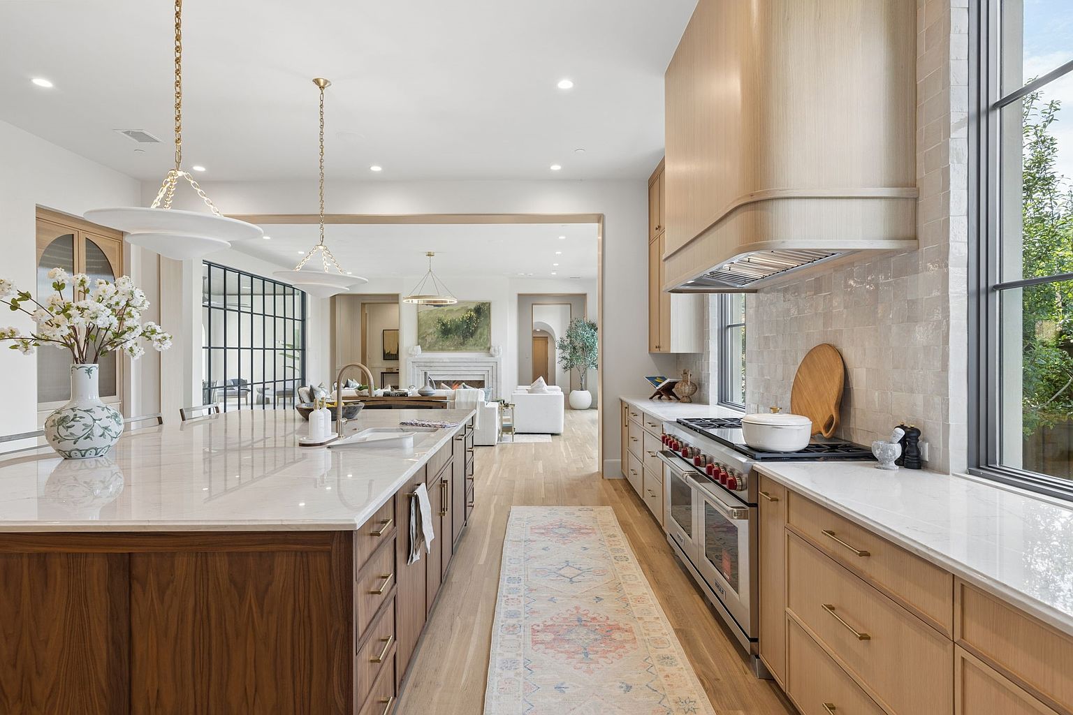 This bright, open-concept kitchen features a large central island with dark wood cabinetry and a light stone countertop, complemented by a long run of light wood cabinets along the wall. A professional-grade range with a custom hood serves as the focal point, while a patterned runner rug adds texture to the light wood flooring. The perspective looks through the kitchen toward a spacious living area, creating an airy and sophisticated atmosphere.