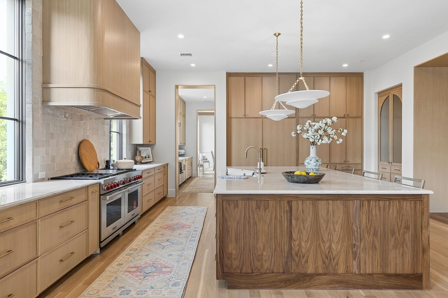 This bright and airy kitchen features a large central island with a dark wood base and a light stone countertop, complemented by light wood cabinetry throughout. A professional-grade range with a custom hood sits against a tiled backsplash, while two modern pendant lights hang above the island. The perspective captures the functional layout, including a view into an adjacent hallway, creating a sophisticated and cohesive design aesthetic.