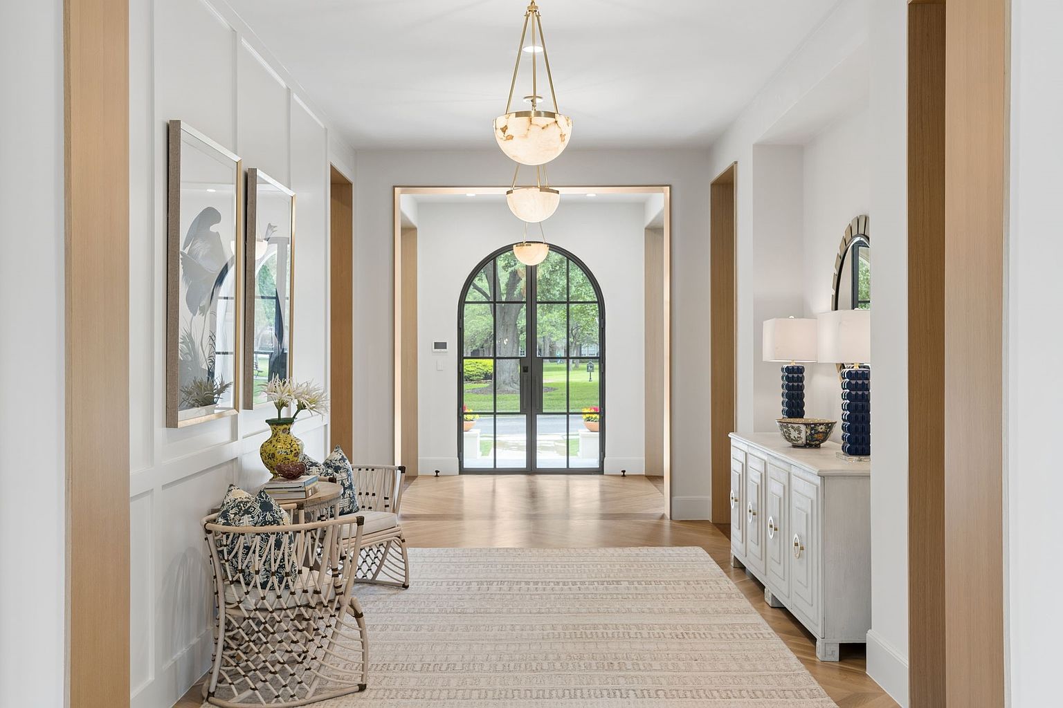 This elegant, light-filled foyer features a long, symmetrical hallway leading to a striking arched glass door that provides a clear view of the outdoors. The space is adorned with white paneled walls, a neutral patterned area rug, and a tiered pendant light fixture that draws the eye upward. To the sides, a console table with matching lamps and a pair of woven chairs create a welcoming and sophisticated atmosphere.