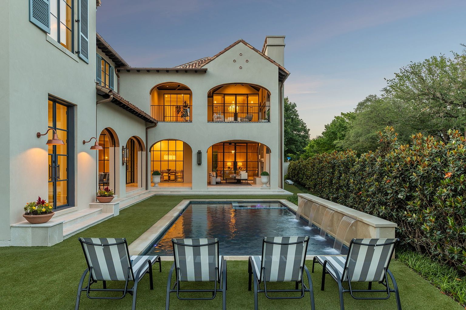 This elegant Mediterranean-style home features a symmetrical rear facade with arched loggias on both the ground and second levels. A rectangular swimming pool with a stone water feature serves as the focal point of the manicured lawn, which is framed by four modern lounge chairs. The warm glow from the interior lighting creates an inviting atmosphere against the soft twilight sky.