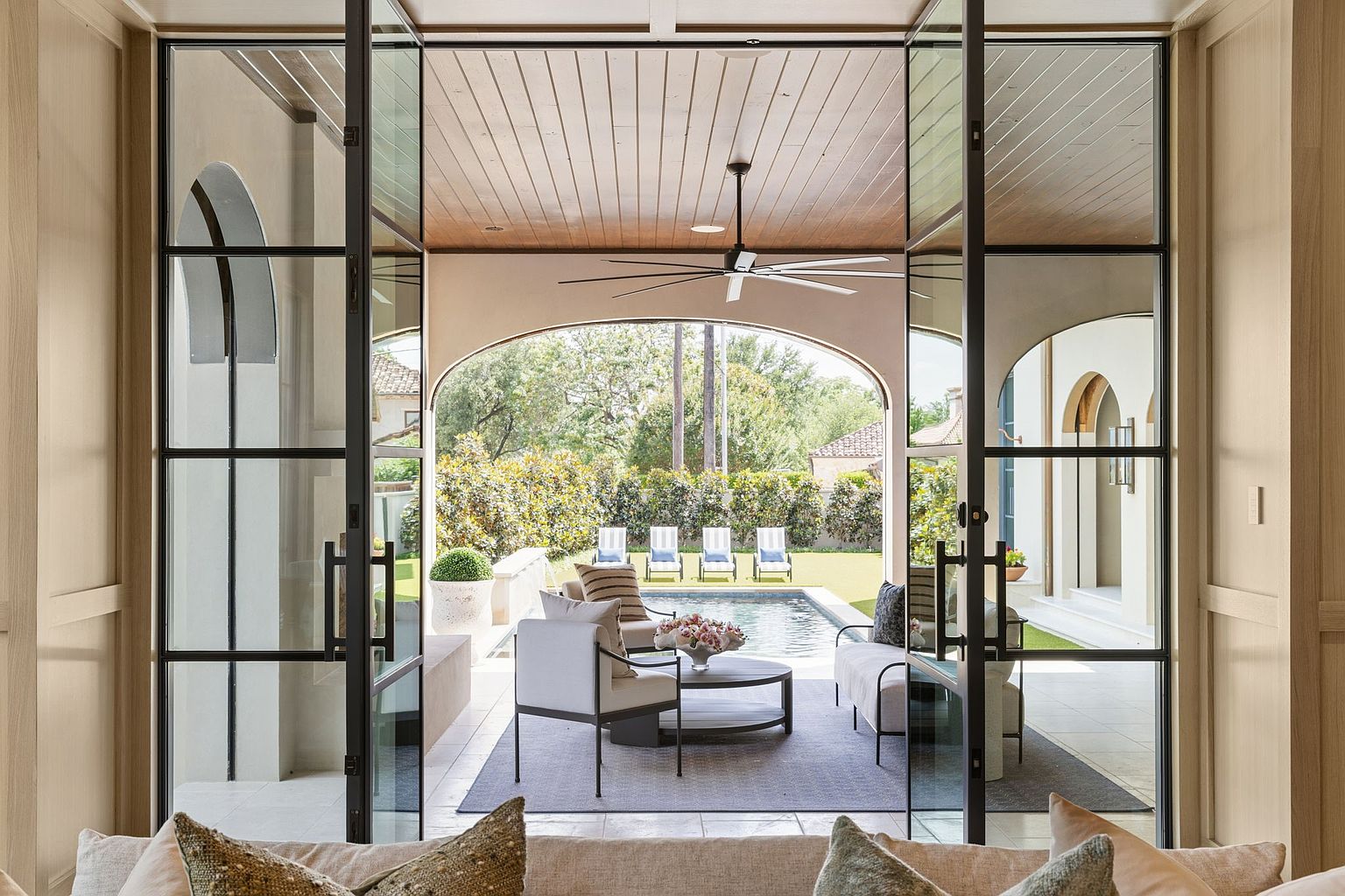 This view from an interior space looks out through large, black-framed glass doors onto a covered patio and pool area. The outdoor living space features a comfortable seating arrangement on a rug, a ceiling fan, and an arched opening that frames the backyard pool and lawn. The scene conveys a seamless transition between indoor and outdoor living, highlighting a sophisticated and airy architectural design.