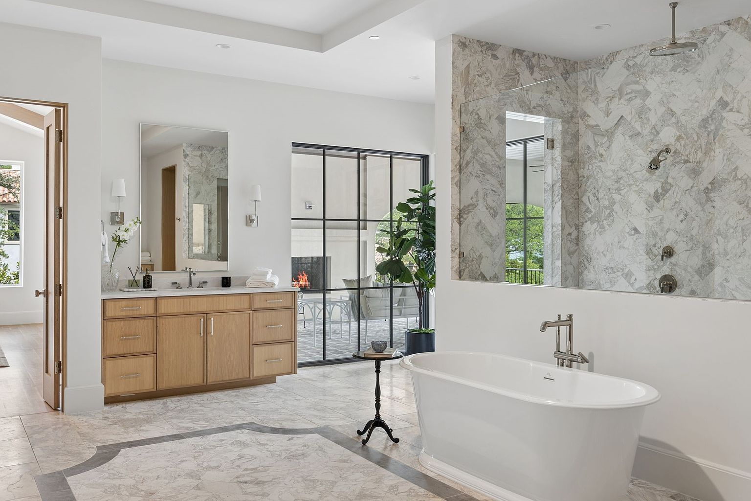 This luxurious primary bathroom features a freestanding soaking tub, a light wood vanity with a rectangular mirror, and a spacious walk-in shower with patterned tile walls. A large glass-paned wall provides a view into an outdoor patio area with a fireplace, creating an airy and connected atmosphere. The room is finished with elegant stone flooring and neutral tones, offering a sophisticated and serene spa-like aesthetic.