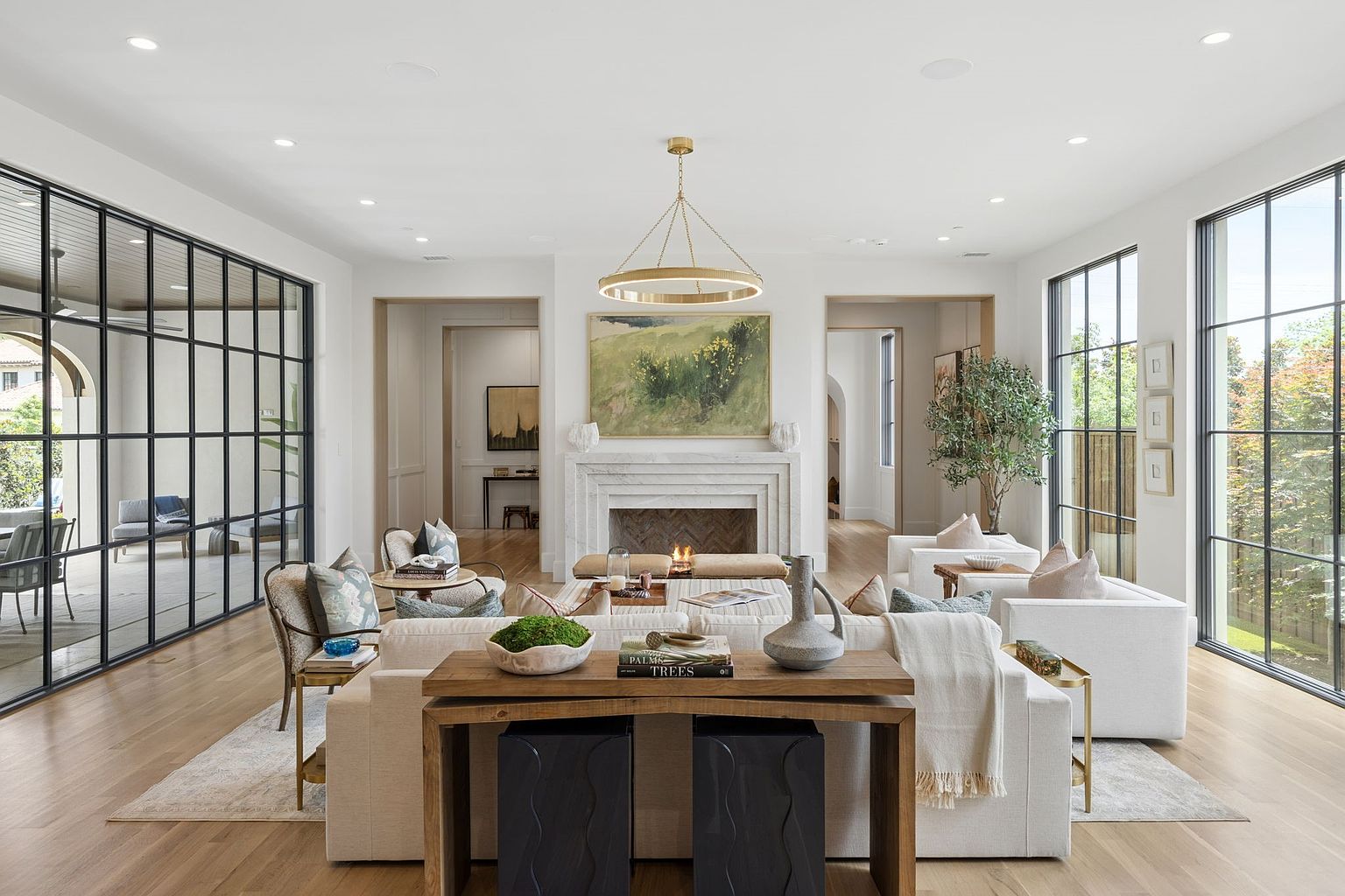 This sophisticated living room features a symmetrical layout centered around a stone-clad fireplace with a large landscape painting above. The space is framed by expansive, black-framed windows that offer views of the outdoors and provide abundant natural light. A neutral color palette, light wood flooring, and a modern gold chandelier create an airy, high-end atmosphere perfect for entertaining.