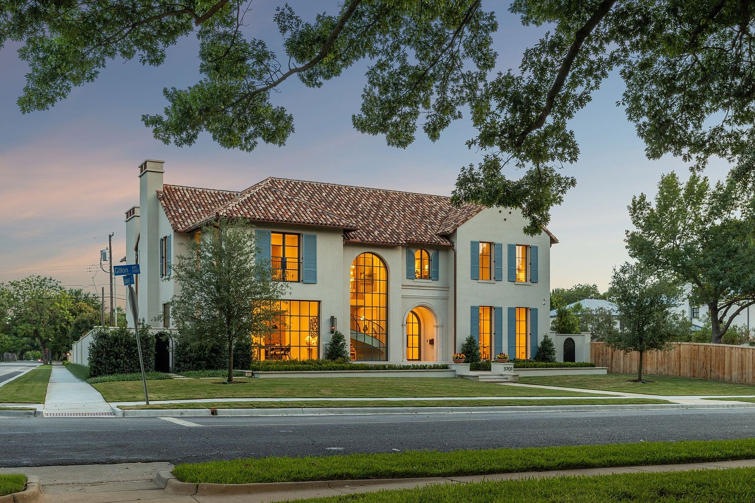This elegant two-story Mediterranean-style residence features a light-colored stucco facade, a classic red-tiled roof, and distinctive blue shutters. The front elevation is highlighted by a grand arched entryway and large, multi-paned windows that allow warm interior light to glow against the twilight sky. Set on a manicured corner lot, the home is framed by mature trees and a neatly landscaped lawn, creating a sophisticated and inviting curb appeal.