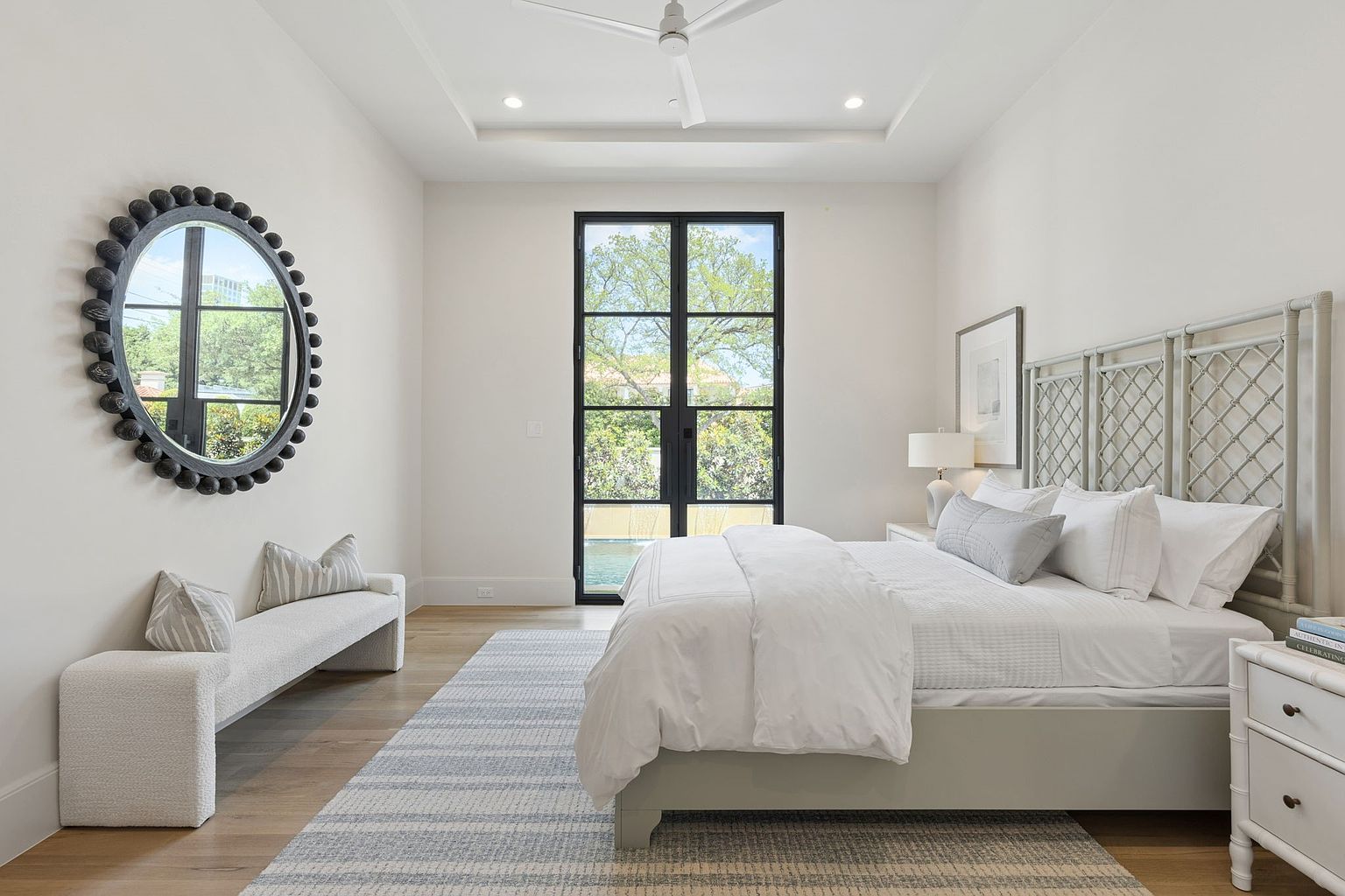 This bright and airy primary bedroom features a neutral color palette, light wood flooring, and a large area rug that anchors the space. A textured, light-colored bed frame serves as the focal point, complemented by a sleek bench placed beneath a large, decorative circular mirror on the wall. Tall, black-framed glass doors provide a view of the outdoors, allowing ample natural light to fill the room.