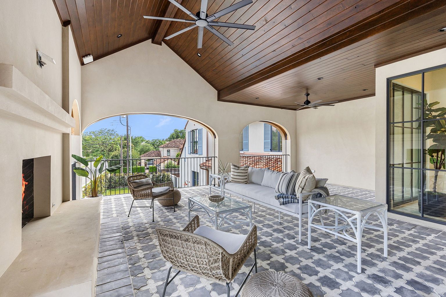 This expansive covered balcony features a sophisticated outdoor living area with a stone fireplace and patterned tile flooring. The space is furnished with a white sofa, glass-topped tables, and woven chairs, all set under a high, wood-paneled vaulted ceiling with integrated fans. Large arched openings provide an open-air feel and offer views of the surrounding neighborhood.