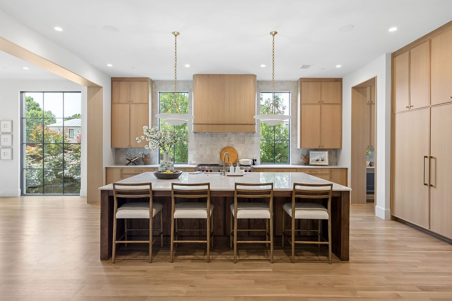This bright and airy kitchen features a large central island with seating for four, complemented by light wood cabinetry and a neutral stone backsplash. The space is anchored by a professional-grade range hood and symmetrical windows that provide ample natural light and views of the outdoors. The design aesthetic is clean and modern, utilizing a warm, cohesive color palette that creates an inviting atmosphere.