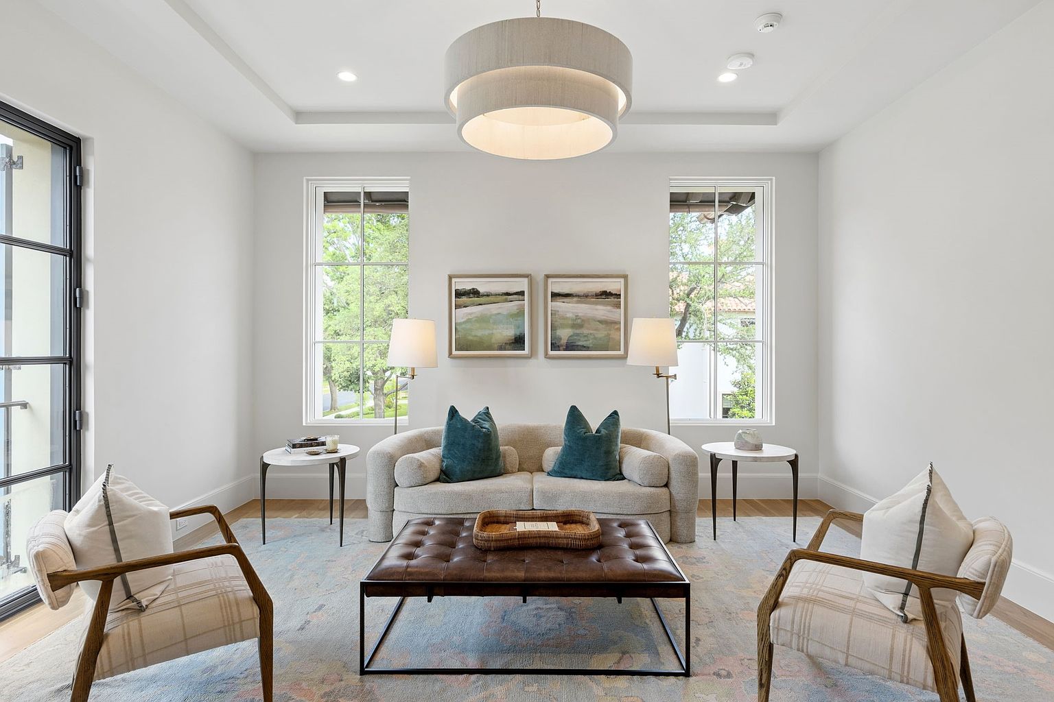 This bright and airy living room features a symmetrical layout centered around a plush, light-toned sofa adorned with teal accent pillows. A dark leather tufted ottoman serves as a coffee table, flanked by two patterned armchairs and matching side tables with elegant lamps. The space is anchored by a large, soft-toned area rug and illuminated by a tiered drum pendant light, creating a sophisticated and inviting atmosphere.