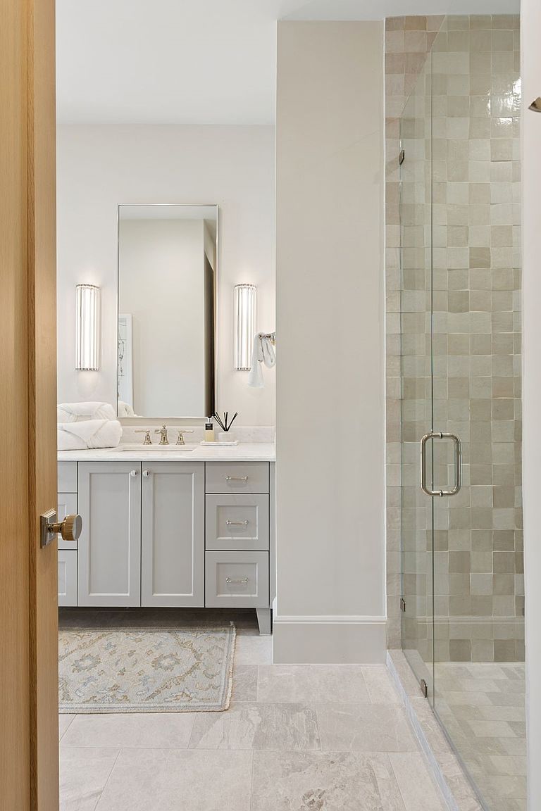 This bright and airy bathroom features a light gray vanity with a stone countertop and a rectangular mirror flanked by vertical wall sconces. To the right, a glass-enclosed shower showcases textured, neutral-toned square tiles that create a subtle, sophisticated pattern. The perspective is captured from a doorway, looking into the space, which is finished with light-colored floor tiles and a decorative area rug.
