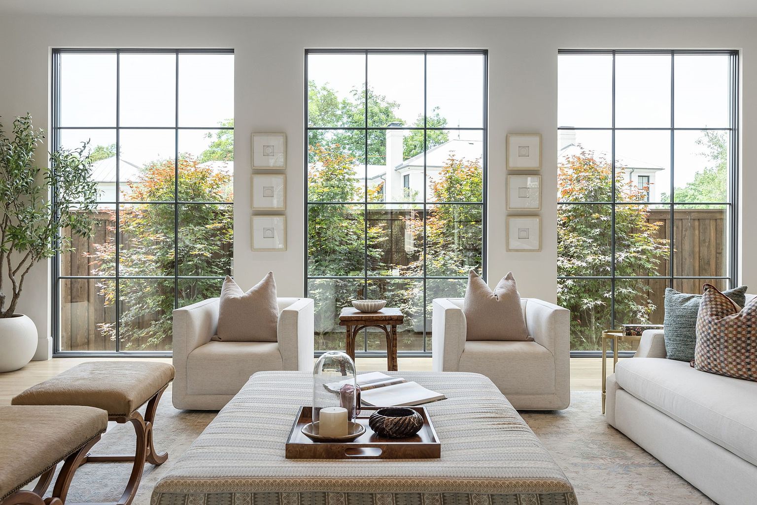 This bright and airy living room features a symmetrical layout centered around a large, fabric-upholstered ottoman and two plush armchairs. Three expansive, grid-paned windows provide abundant natural light and a clear view of the lush greenery outside. The space is decorated with neutral tones, framed wall art, and a potted tree, creating a sophisticated and tranquil atmosphere.