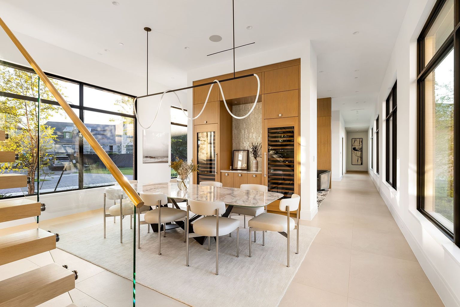 This is a bright and modern dining room featuring a marble-topped table surrounded by six chairs with metal legs. A unique, linear light fixture hangs above the table, and a large window provides ample natural light. The room also includes built-in wine storage and a hallway with windows, creating an open and airy atmosphere.