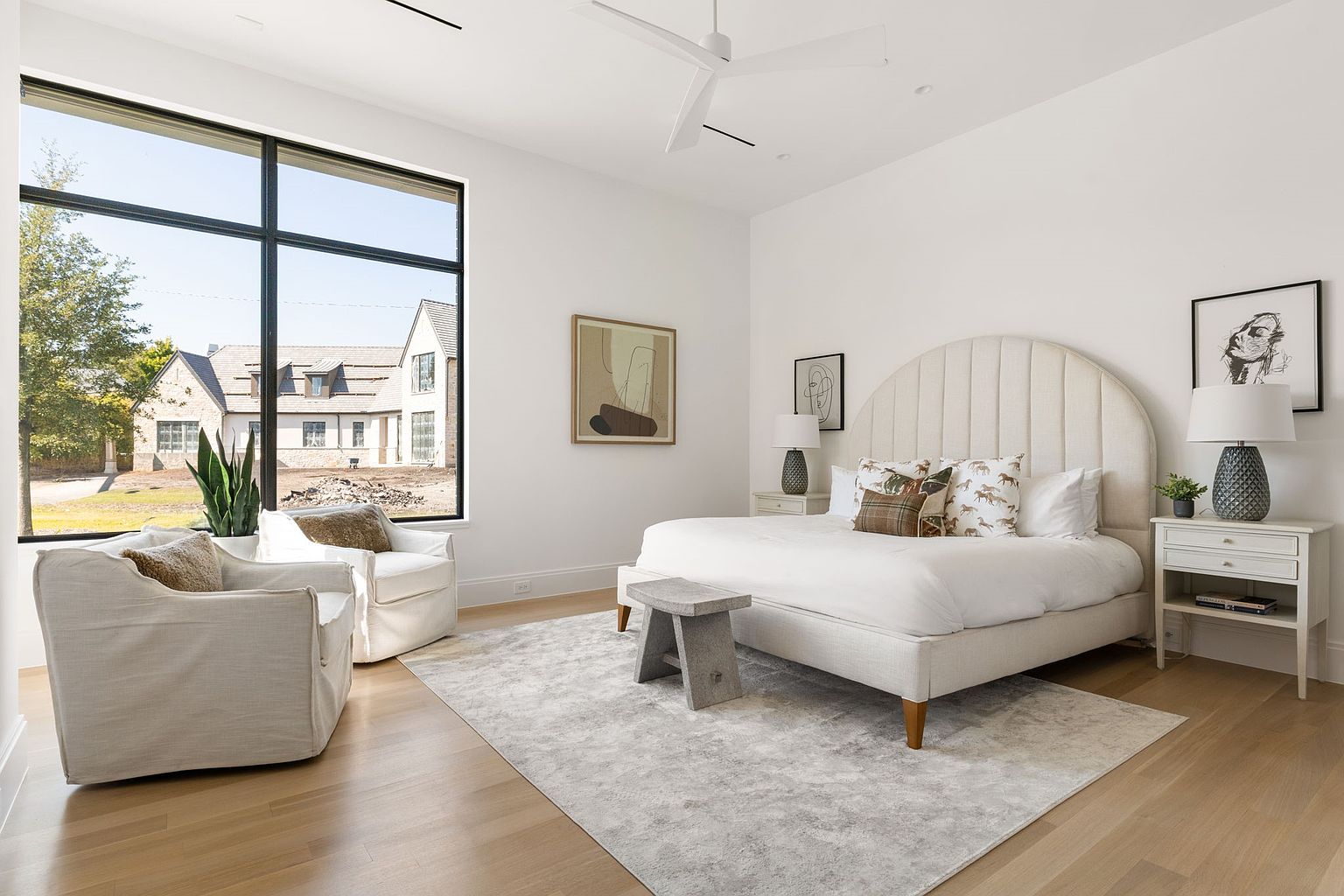 This is a bright and airy primary bedroom featuring a large window with a view of the exterior. The room is decorated in neutral tones with a large upholstered headboard, white bedding, and light wood floors. Two armchairs sit near the window, and a gray rug anchors the space, creating a serene and inviting atmosphere.