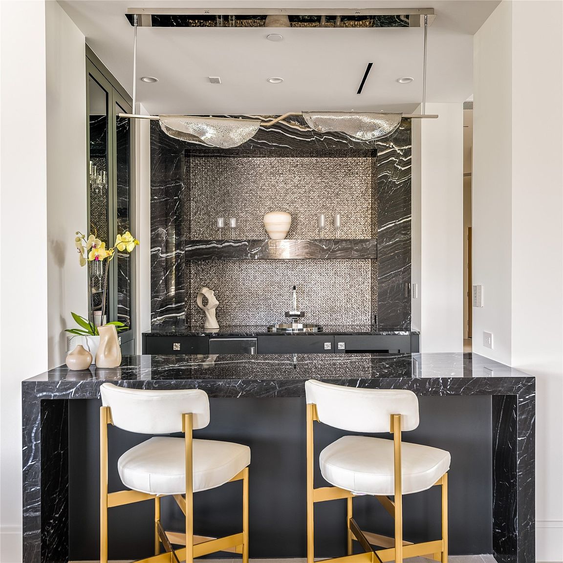 This image showcases a stylish dining area featuring a black marble countertop bar with two white and gold bar stools. The backdrop includes a mosaic tile wall with a shelf displaying decorative items, complemented by a modern chandelier. The overall aesthetic is luxurious and contemporary, perfect for entertaining.