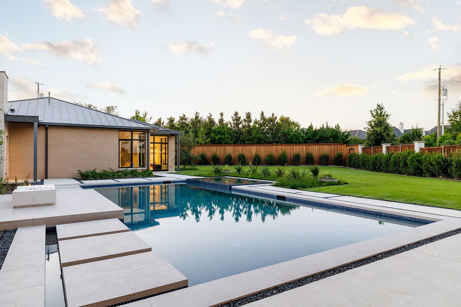 This image showcases a modern backyard featuring a rectangular swimming pool with integrated spa elements. The pool is surrounded by light-colored stone pavers and dark gravel accents, leading to a contemporary brick house with large windows. Lush green lawn and manicured hedges create a serene and private outdoor space.