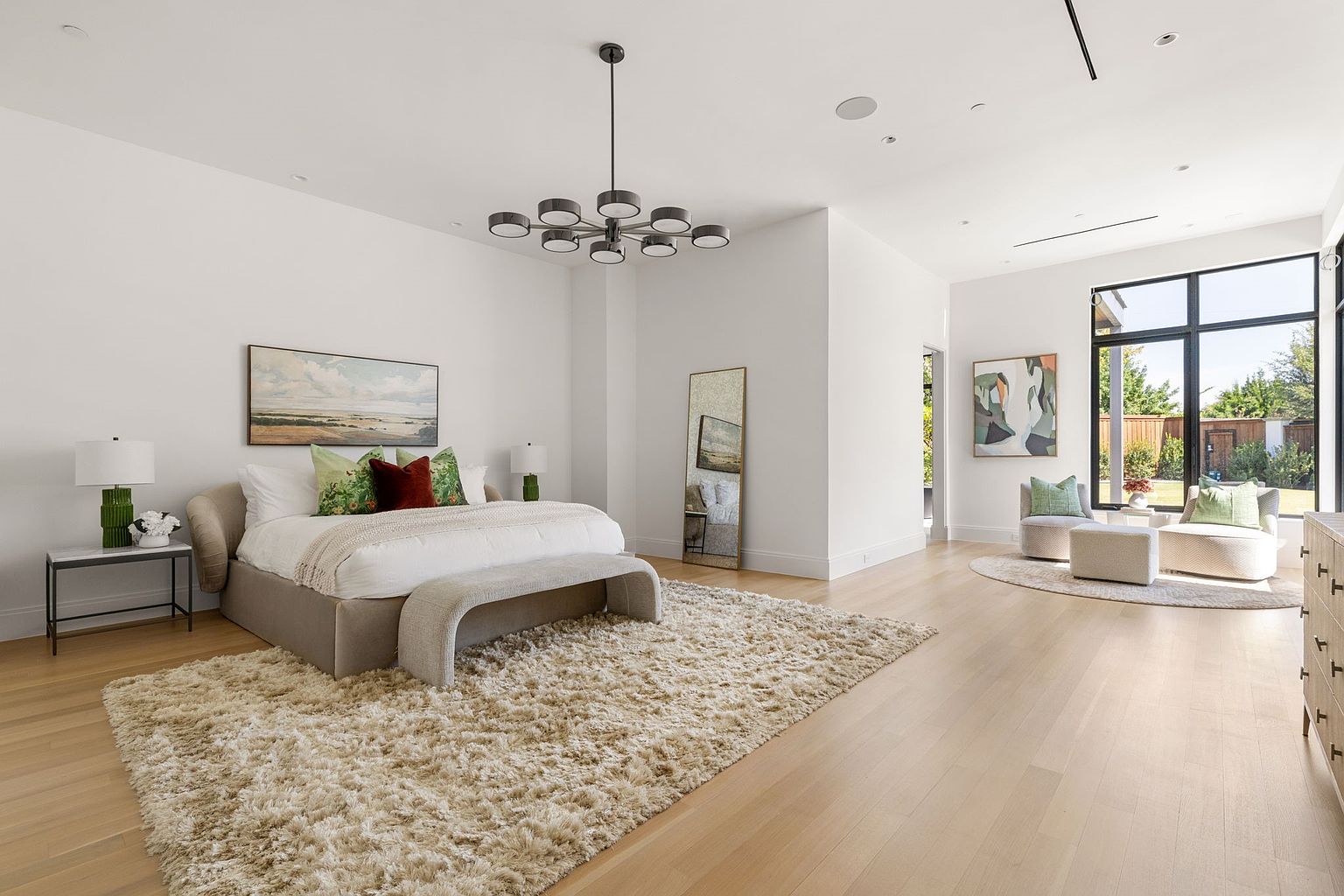 This is a spacious primary bedroom featuring a large bed with neutral bedding and decorative pillows, complemented by a bench at the foot of the bed. A modern chandelier hangs from the ceiling, and a full-length mirror stands against the wall. The room has a light and airy feel, enhanced by the light wood flooring and large windows that provide ample natural light.