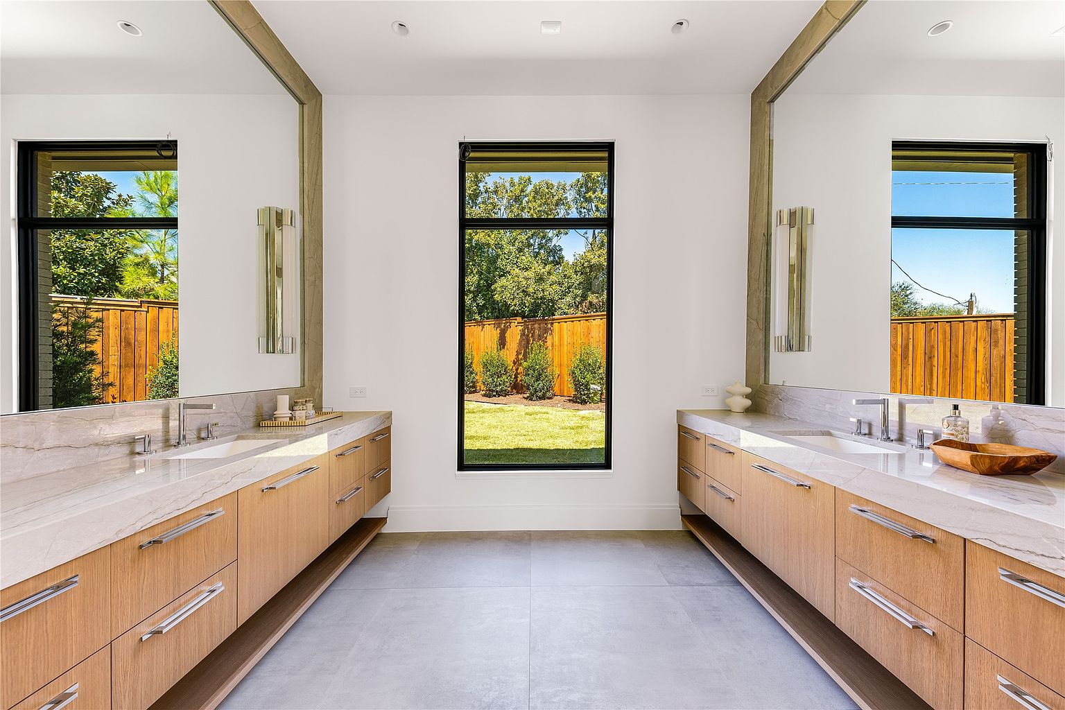 This is a bright and modern primary bathroom featuring dual vanities with light wood cabinetry and marble countertops. Large mirrors are framed with a natural wood accent, and a tall window provides natural light and a view of the backyard. The neutral color palette and clean lines create a spa-like atmosphere.