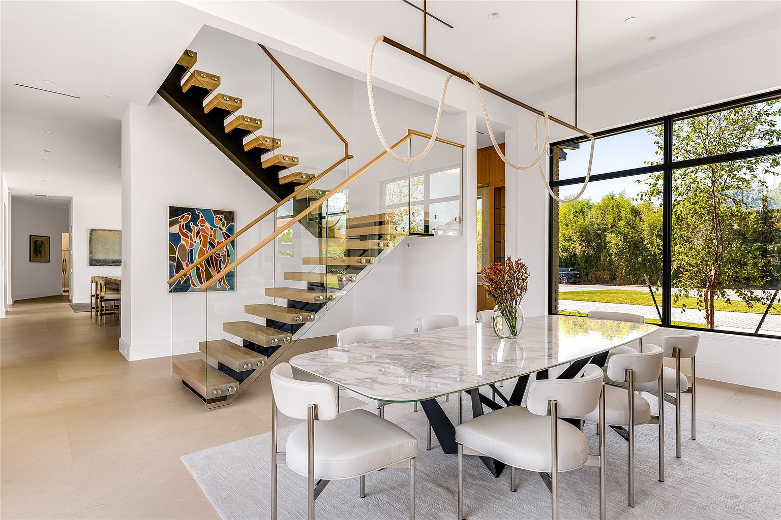 This interior shot showcases a modern dining room with a marble-topped table surrounded by stylish white chairs with silver legs. A unique chandelier hangs above the table, and a large window provides natural light and a view of the outdoors. The room also features a contemporary staircase with glass railings and wooden steps, adding an architectural element to the space.