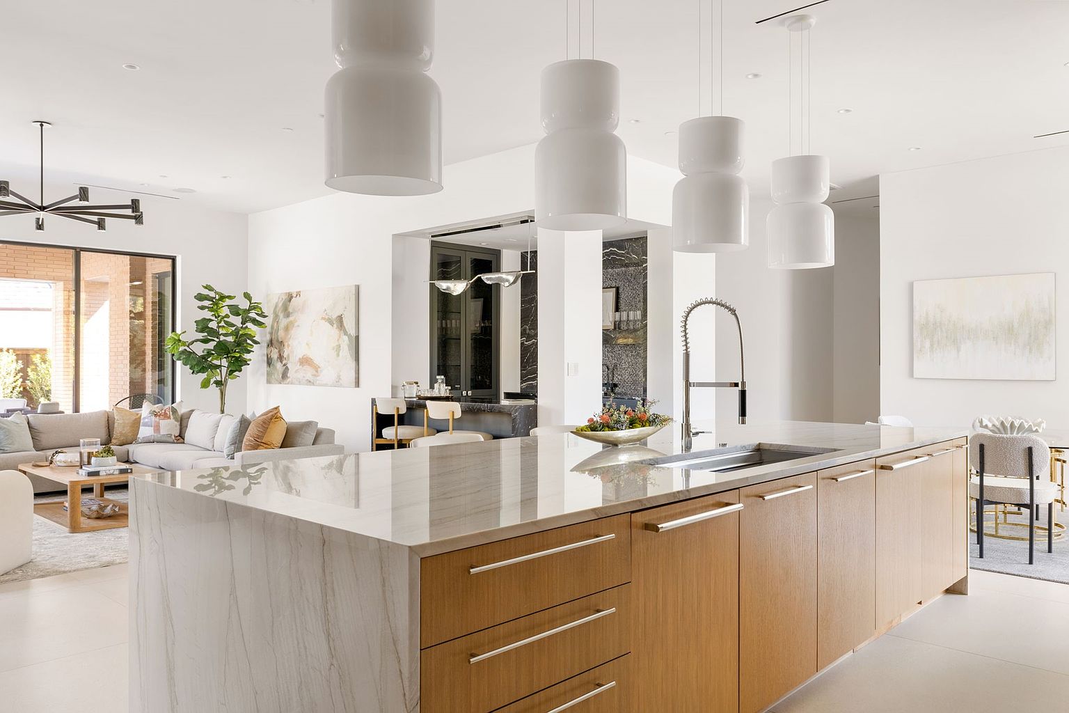 This is a bright and airy kitchen featuring a large island with a marble countertop and wooden cabinetry. The open-concept design seamlessly connects the kitchen to the living area, enhancing the sense of space. Modern pendant lights hang above the island, adding a touch of elegance to the contemporary design.