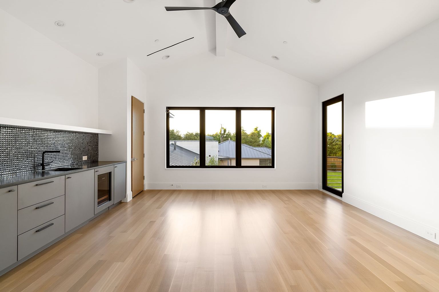 This is a bright and modern living room featuring light hardwood floors and white walls, creating a spacious and airy feel. A sleek wet bar with gray cabinetry and a dark backsplash is situated on one side, while a large window and a glass door provide ample natural light and views of the outdoors. The room is minimalist yet stylish, perfect for entertaining or relaxing.