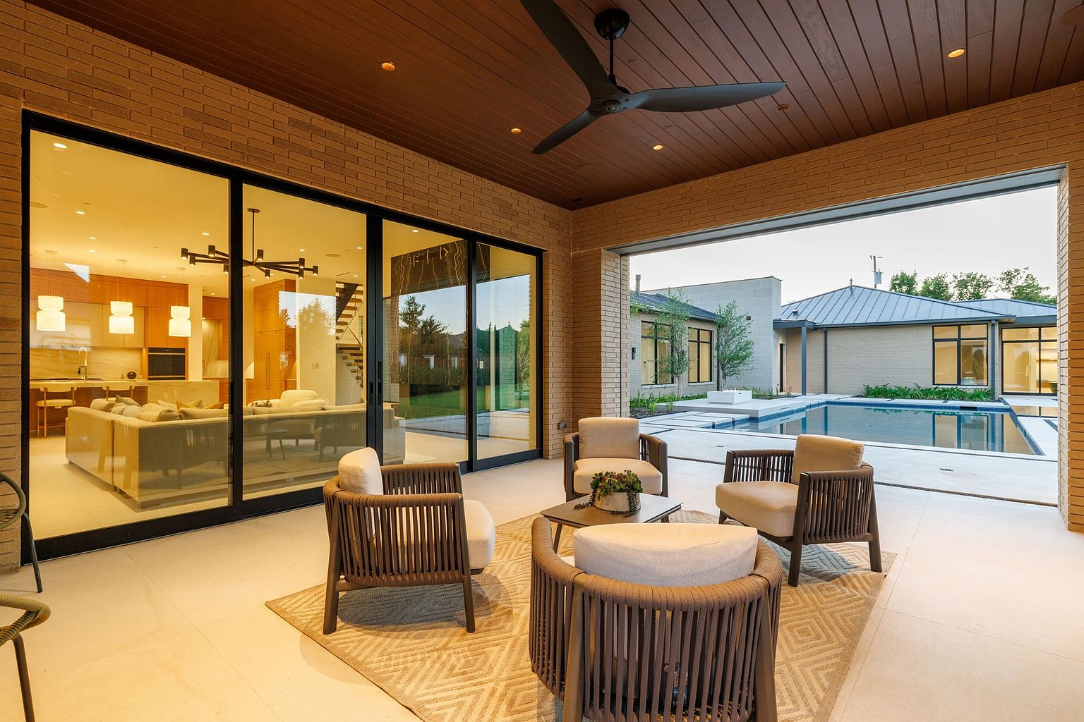 This image showcases a luxurious outdoor patio area, featuring comfortable lounge seating arranged around a central coffee table, set upon a patterned rug. The patio is partially enclosed with brick walls and a wood-paneled ceiling with a ceiling fan, while large sliding glass doors offer views into the home and out to a modern pool area and backyard. The space exudes a sense of relaxation and upscale outdoor living.