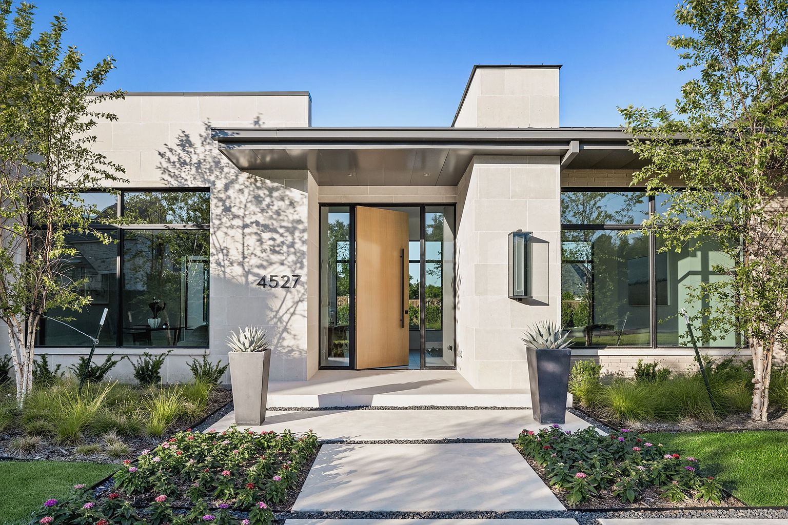 This is a striking front exterior view of a modern home, emphasizing the entryway. The house features a light-colored stone facade, large windows, and a prominent wooden front door that is slightly ajar, inviting viewers in. The landscaping is meticulously designed with a mix of greenery, flowers, and decorative gravel, enhancing the property's curb appeal.