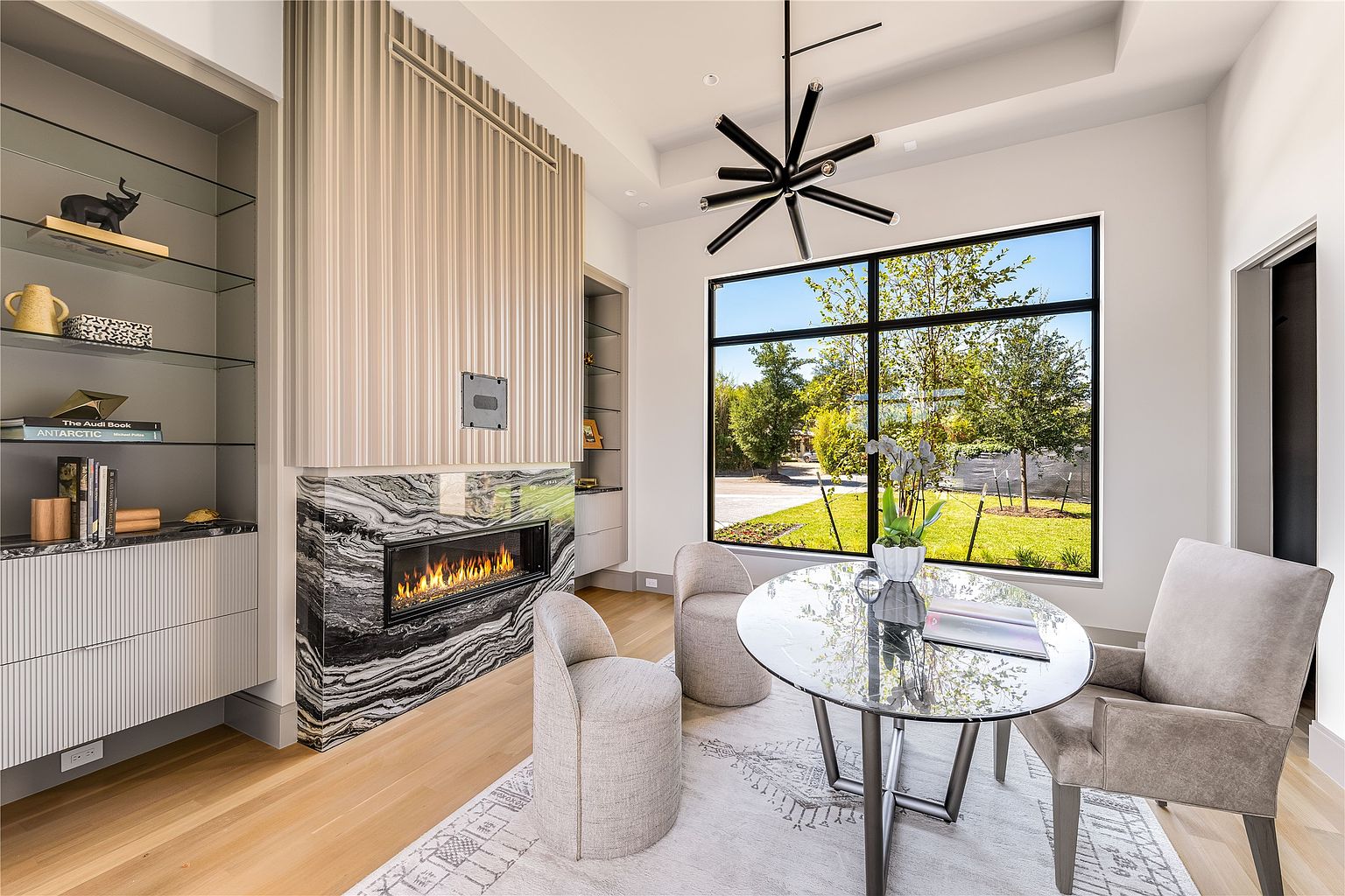 This interior shot showcases a modern office space featuring a striking fireplace with a black and white marble surround and a built-in shelving unit. A round glass table is surrounded by three upholstered chairs, positioned in front of a large window that offers a view of the exterior landscape. The room is illuminated by a contemporary chandelier, creating a sophisticated and inviting atmosphere.