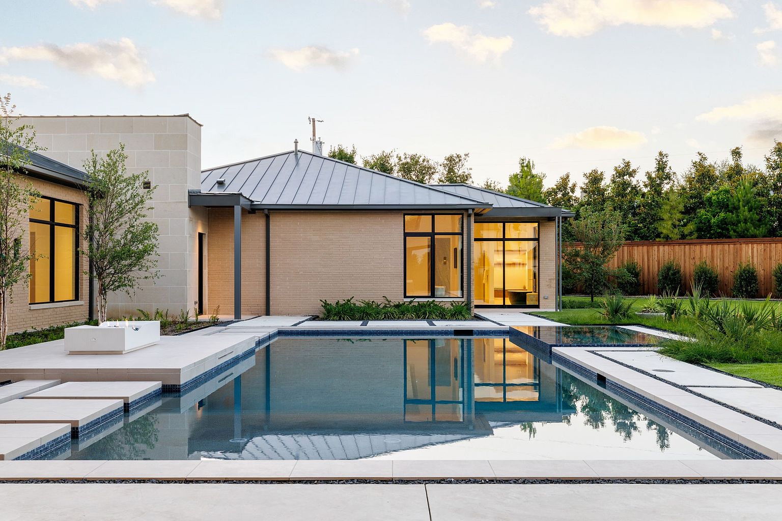 This image showcases a modern home's exterior with a stunning pool and spa area. The pool features clean lines and is surrounded by a light-colored patio, complemented by lush landscaping and a wooden fence in the background. The architecture of the house is contemporary, with large windows reflecting the pool's water, creating a serene and luxurious atmosphere.