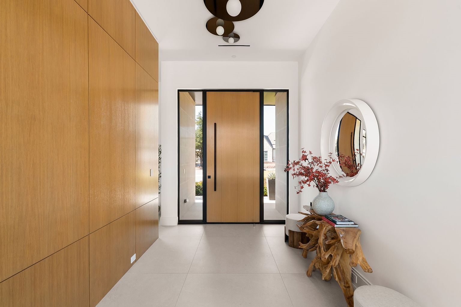 This is an interior shot of a modern hallway featuring a large wooden door with black trim and a long black handle, complemented by a light wood paneled wall on the left. A round mirror hangs on the white wall to the right, above a unique wooden console table adorned with a vase of red flowers and books. The hallway is well-lit with modern ceiling fixtures and natural light streaming through the door, creating a bright and inviting space.
