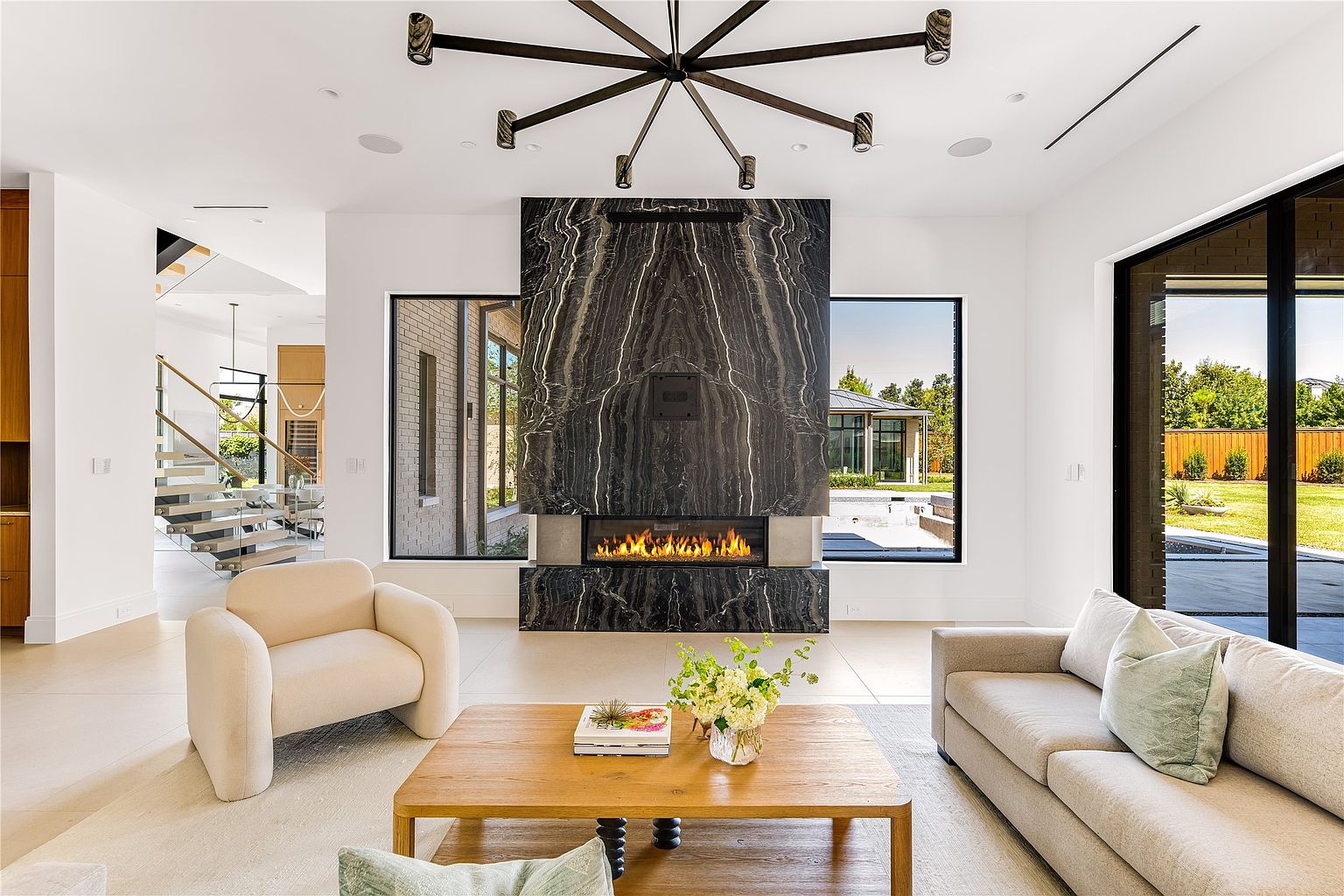 This is a bright and modern living room featuring a striking black marble fireplace as a focal point. The room is furnished with a beige sofa, a matching armchair, and a wooden coffee table adorned with flowers and books. Large windows offer views of the exterior, enhancing the open and airy feel of the space.