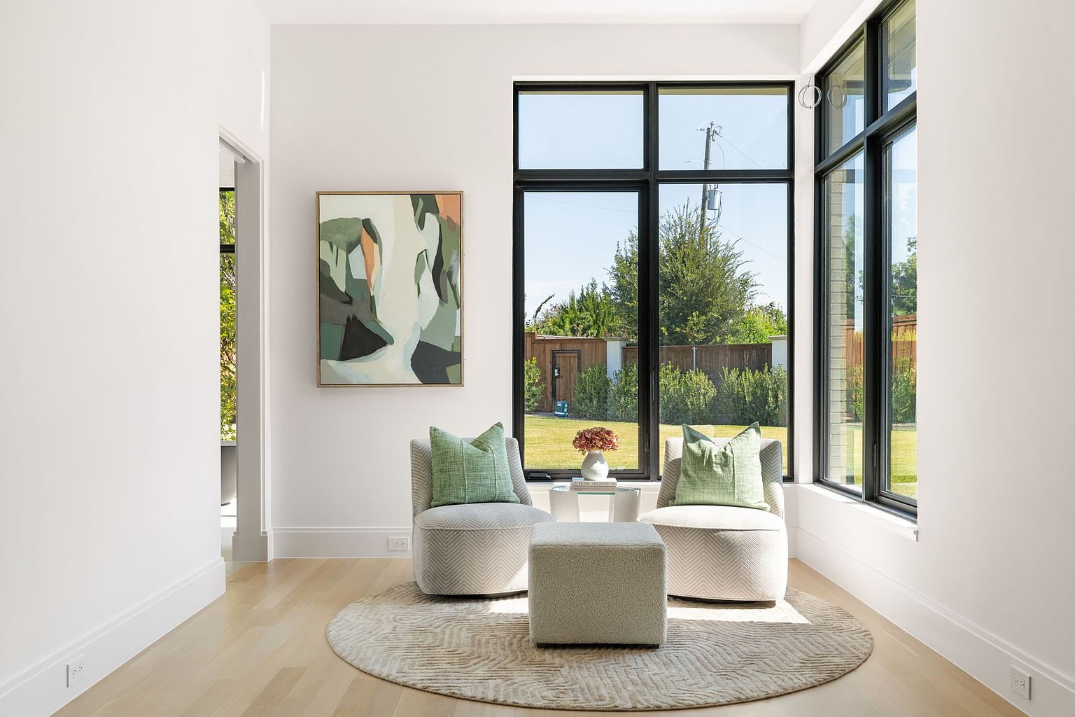 This sunroom features two armchairs with green pillows, a square ottoman, and a round rug. A large window provides ample natural light and a view of the backyard. The room is decorated with an abstract painting and a small table with flowers, creating a cozy and inviting atmosphere.