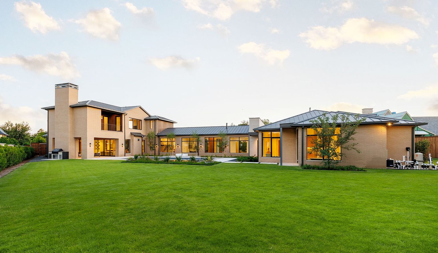This is a rear view of a modern, sprawling home with a well-manicured lawn. The house features a light brick exterior, dark metal roofing, and large windows that allow ample natural light. The design emphasizes clean lines and a seamless integration with the outdoor space, creating a luxurious and inviting atmosphere.