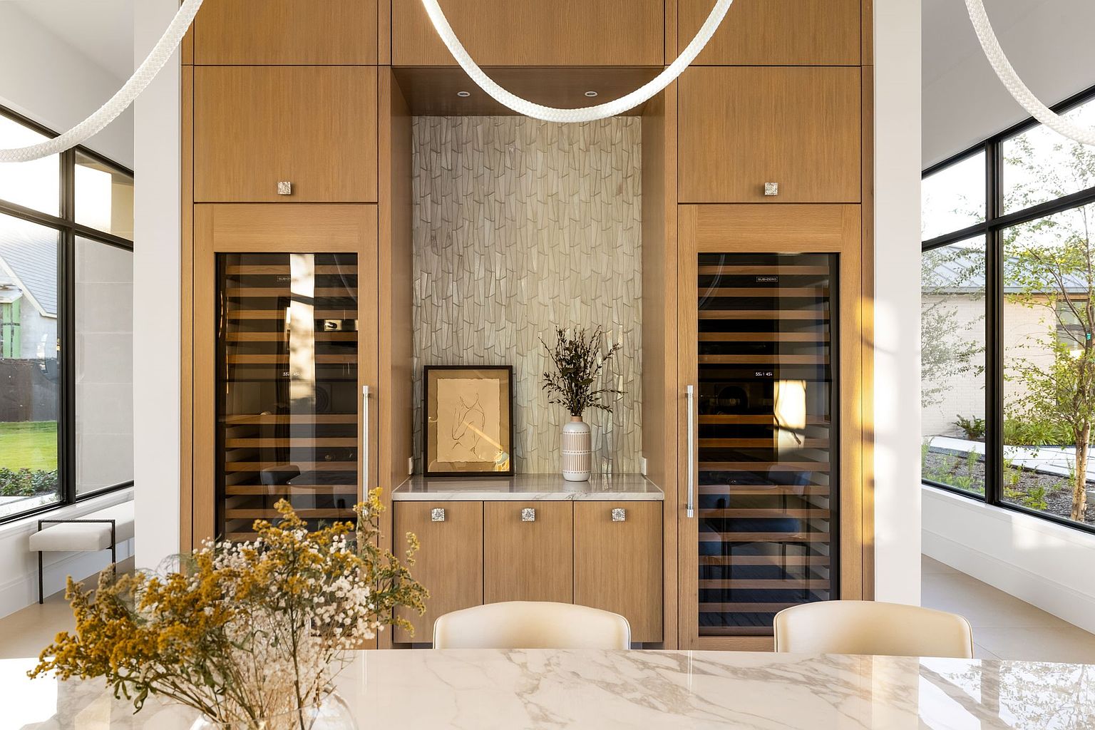 This interior shot showcases a dining area with a built-in cabinet featuring wine coolers on either side of a decorative tiled section. The cabinet is made of light wood and has modern hardware. A marble-topped table with chairs is in the foreground, and large windows provide natural light, creating a bright and inviting space.