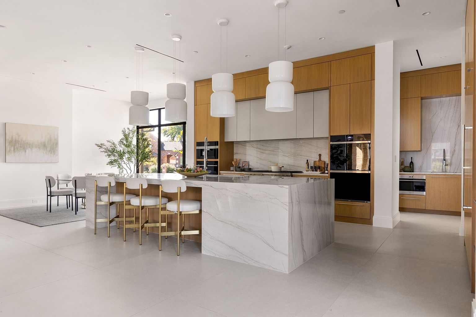 This is a bright and modern kitchen showcasing an expansive island with marble countertops and gold accent bar stools. The kitchen features sleek, light wood cabinetry contrasted by light grey wall cabinets and a marble backsplash. The space is illuminated by pendant lighting, creating an inviting atmosphere, ideal for both cooking and entertaining.