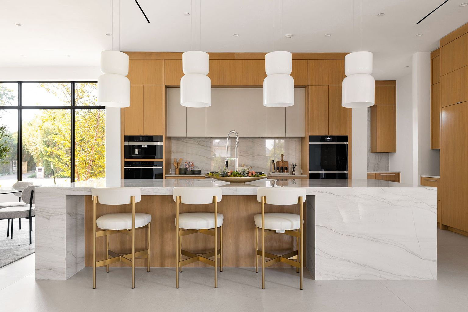 This is a bright and modern kitchen featuring light wood cabinetry and a large marble island with seating. The island is adorned with a decorative bowl, and pendant lights hang above it. Built-in appliances and a large window add to the luxurious feel of the space, creating an inviting atmosphere.