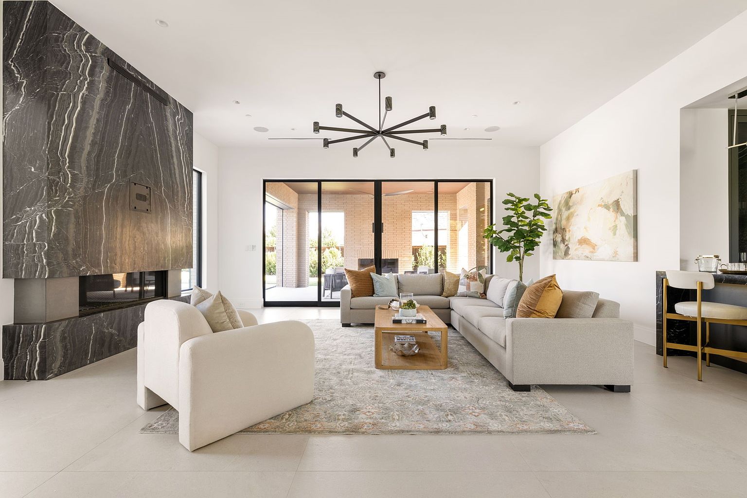 This is a bright and modern living room featuring a large marble fireplace, a neutral-toned sectional sofa, and a unique starburst chandelier. The room opens to an outdoor patio through large sliding glass doors, providing ample natural light. An area rug anchors the seating arrangement, and a piece of abstract art adds a touch of color to the space.