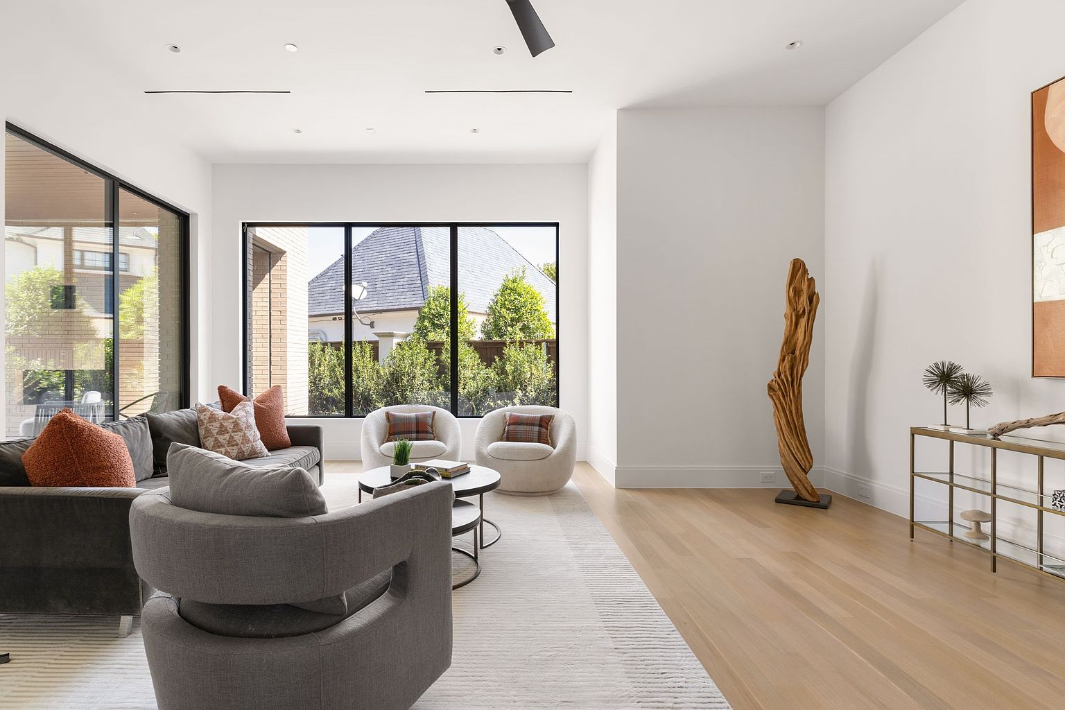 This is a bright and airy living room featuring a modern design with large windows that offer a view of the exterior. The room is furnished with a gray sectional sofa, a round coffee table, and two accent chairs. A light-colored rug anchors the space, and a decorative wooden sculpture adds an artistic touch.