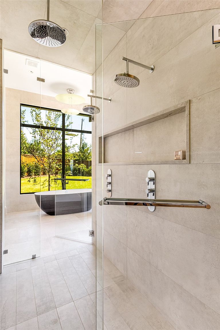 This is a luxurious primary bathroom featuring a glass-enclosed shower with dual shower heads and a built-in niche. The bathroom has large format tiles, a sleek towel bar, and a modern freestanding bathtub visible through the glass. The design emphasizes clean lines and a spa-like atmosphere, enhanced by the natural light from the window overlooking the yard.