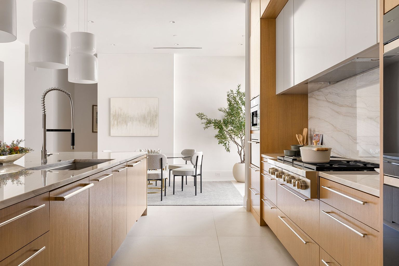 This is a bright and modern kitchen featuring light wood cabinetry with sleek hardware and white upper cabinets. The kitchen is open to a dining area, creating a seamless flow. A large island with a stainless steel sink and a high-end range with a marble backsplash add to the luxurious feel of the space.